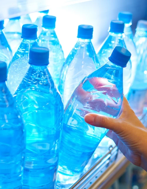Close-up of transparent water bottles with blue caps lined on shelf, with a hand reaching to select one bottle