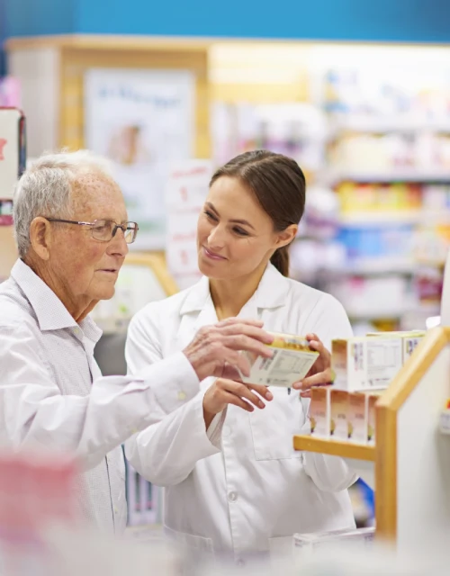 Pharmacy professional consulting with older shopper about medication in bright, well-stocked store aisle.