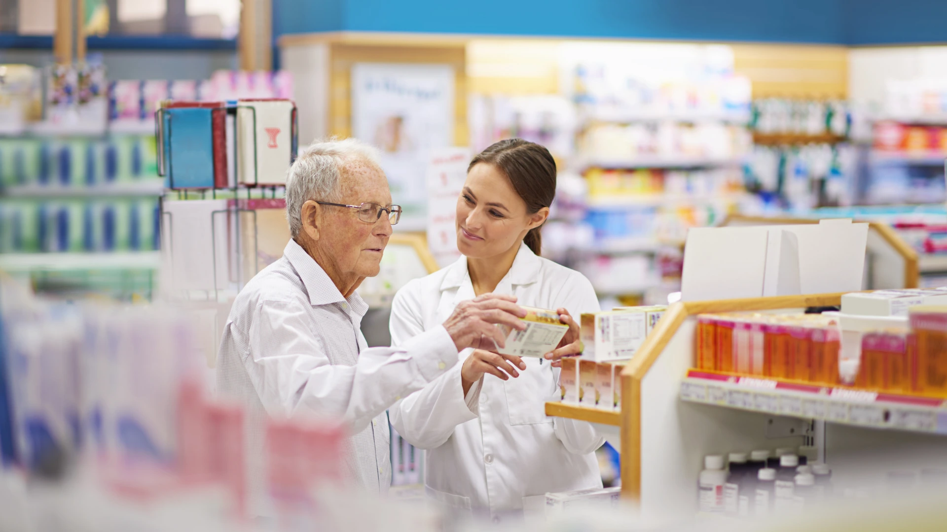Pharmacy professional consulting with older shopper about medication in bright, well-stocked store aisle.