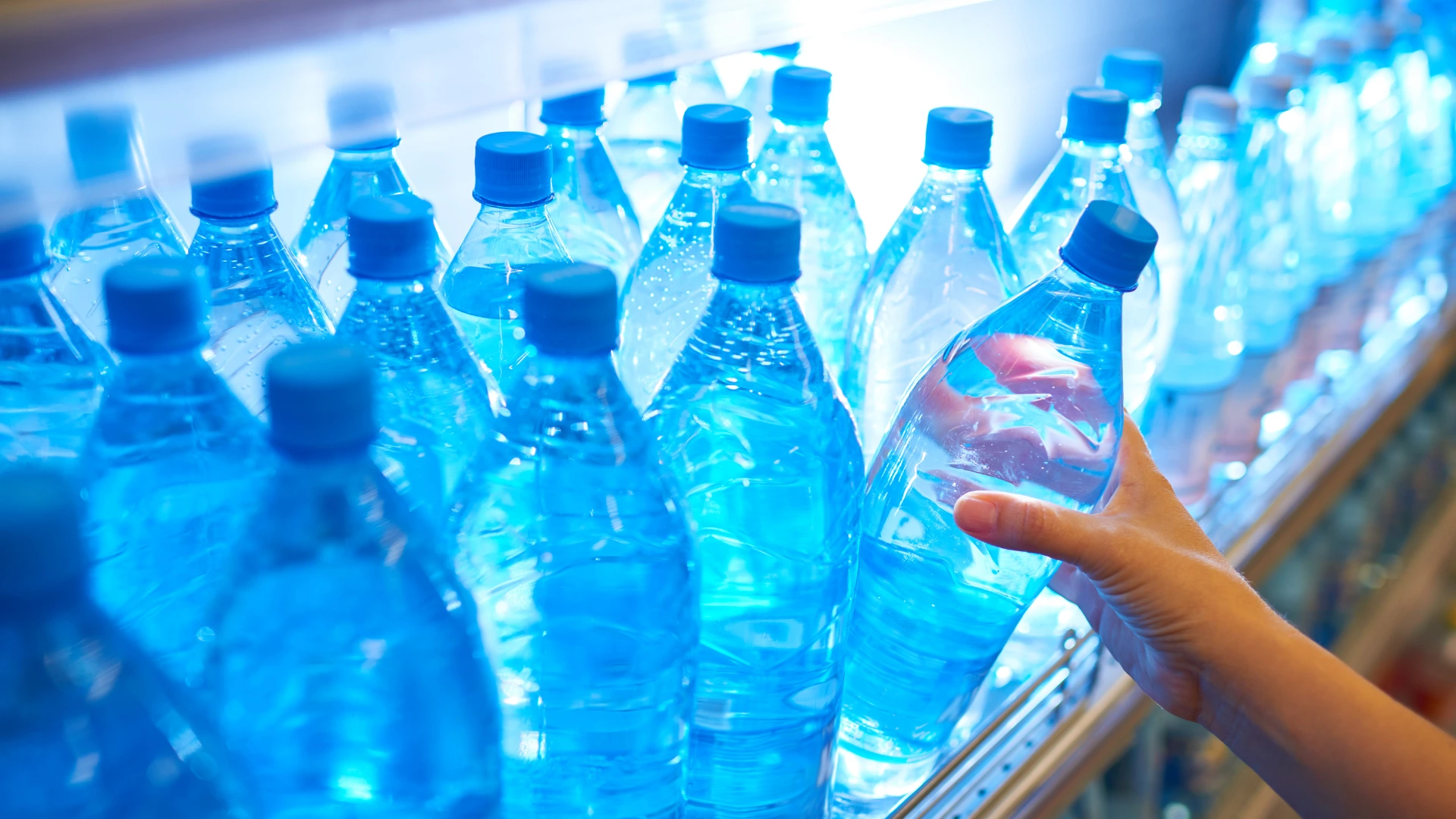 Close-up of transparent water bottles with blue caps lined on shelf, with a hand reaching to select one bottle