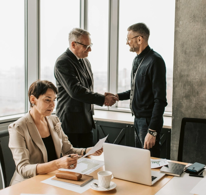Two colleagues shaking hands while another reviews documents at a conference table.