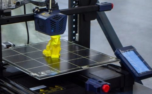 3D printer building a yellow geometric model on heated print bed.