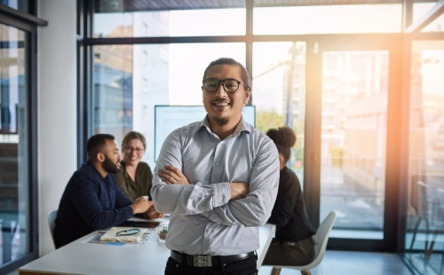 Business professional stands arms crossed, colleagues having meeting at glass office table