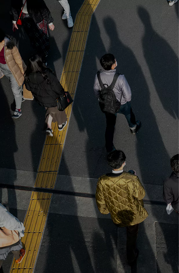 Urban crowd casting elongated shadows while walking along yellow tactile paving on sunlit street.