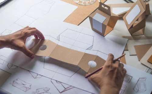 Hands creating cardboard packaging prototype over blueprints and design drawings
